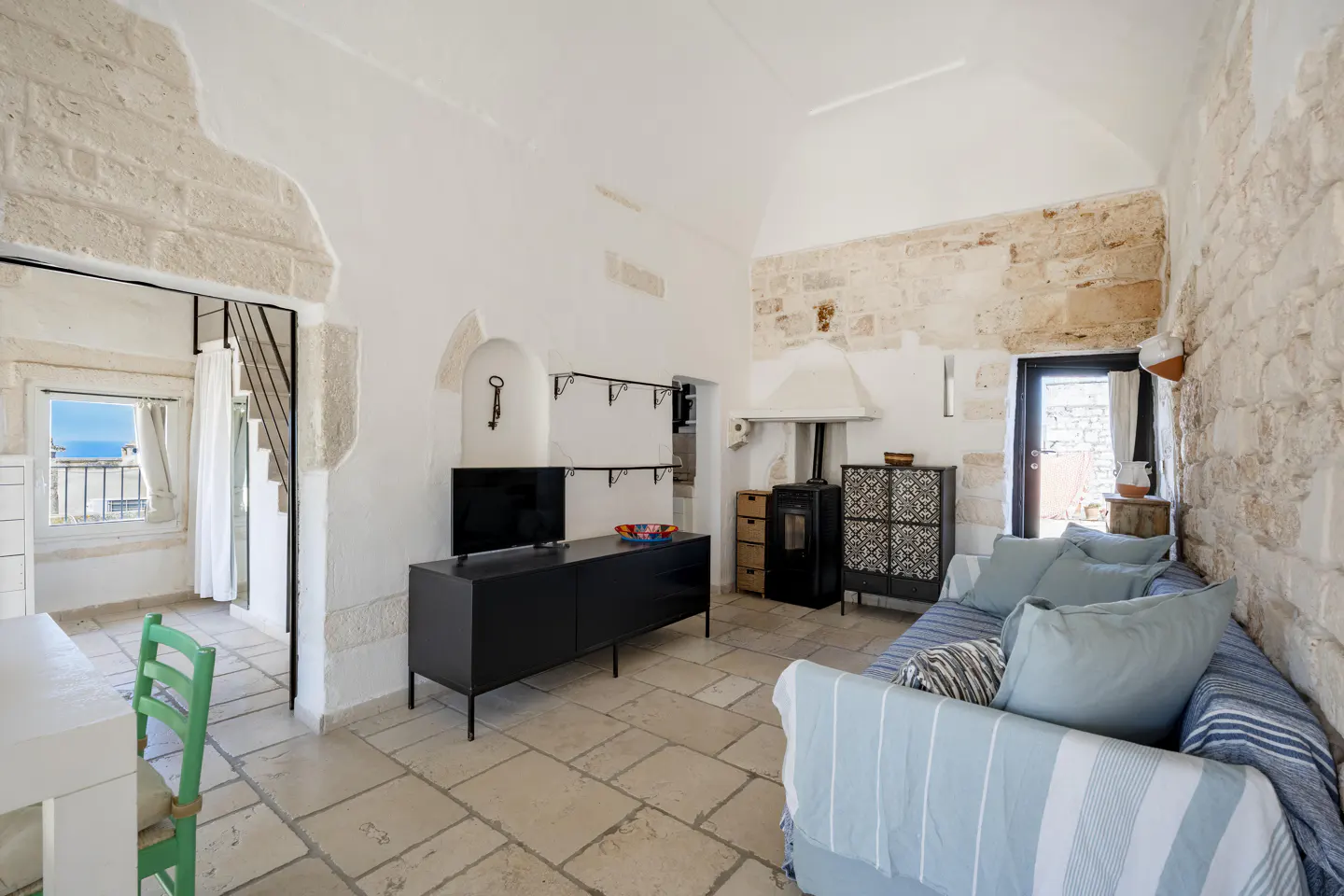 Bright living room with stone walls, tile floor, and vaulted ceiling. A black TV stand and wood-burning stove are visible. A blue sofa sits near a doorway.