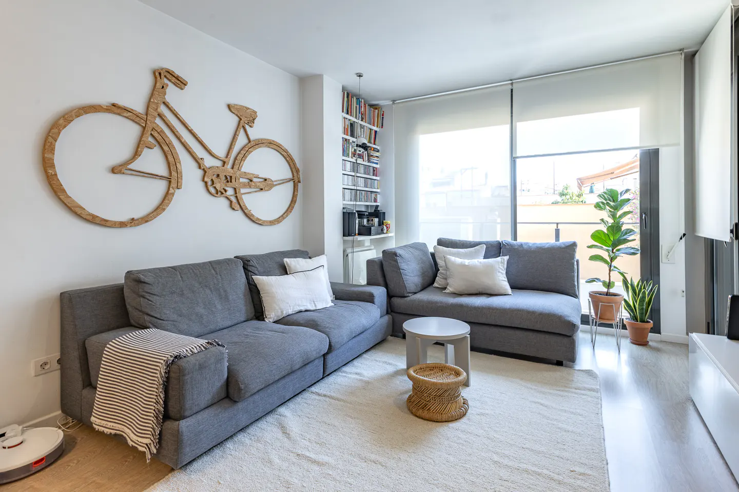 Living room with gray sofas, white pillows, and a wooden bicycle wall decoration. A white rug covers the floor.