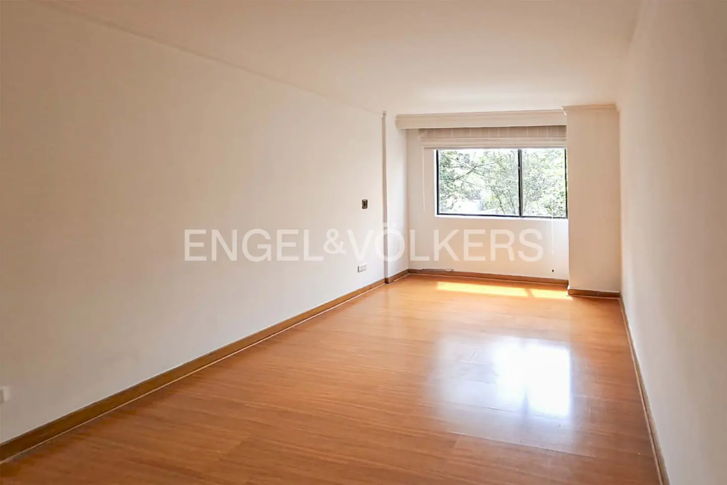 Empty room with light wood floors, white walls, and a window showing trees outside. Engel & Volkers logo on the wall.