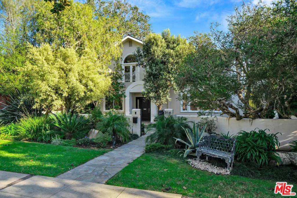 Two-story beige home with a stone walkway, lush landscaping, and a decorative metal bench in the front yard.