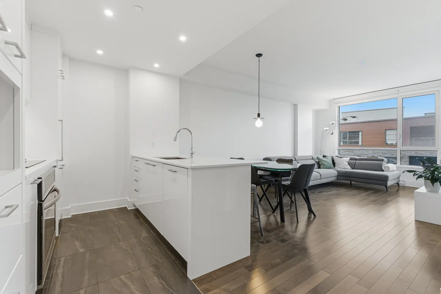 Bright, modern apartment interior with white kitchen island, black chairs, and gray sectional sofa near a large window.