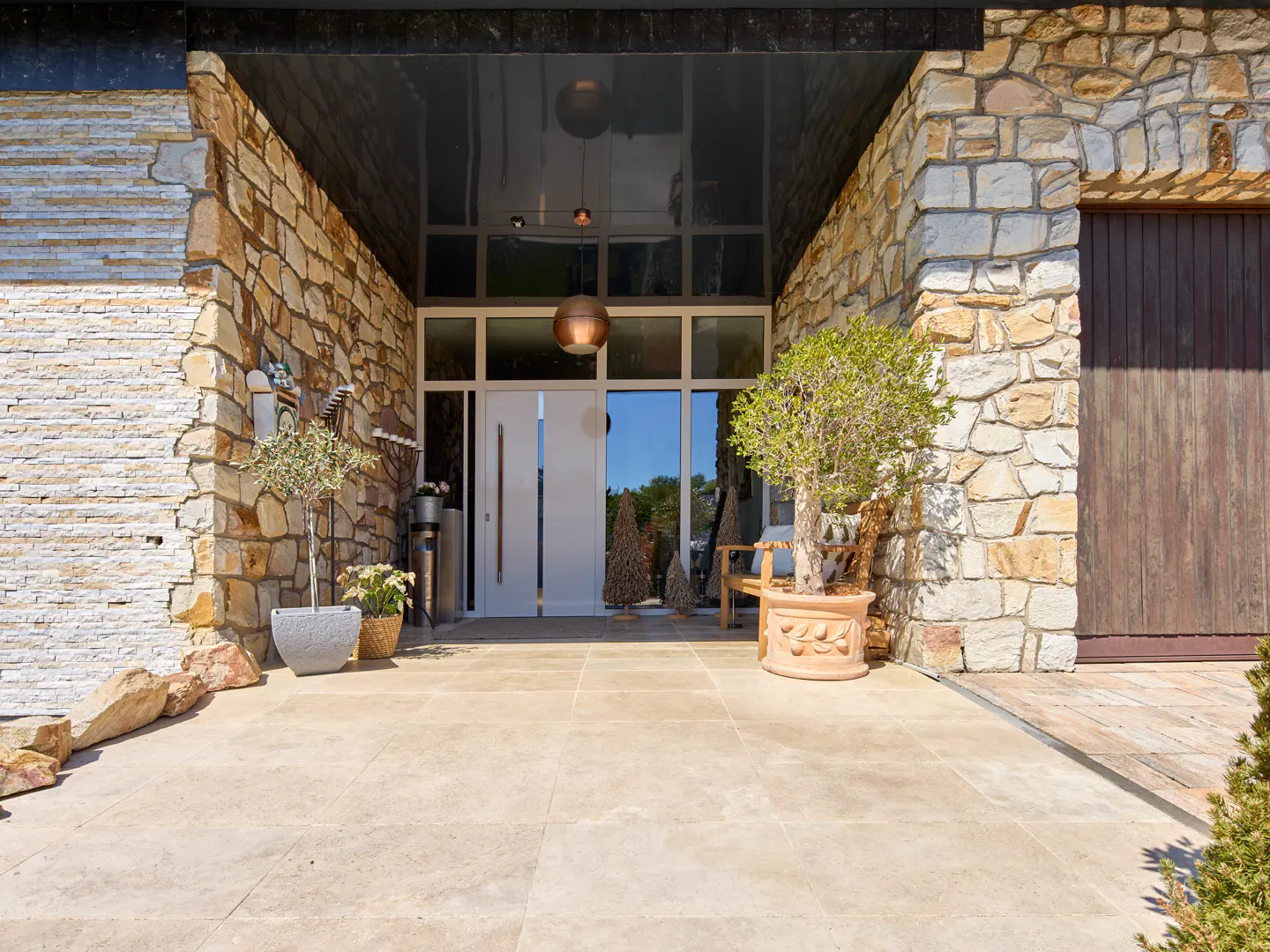 Stone facade of a modern home with a white front door, large windows, and potted plants on a beige tiled patio.