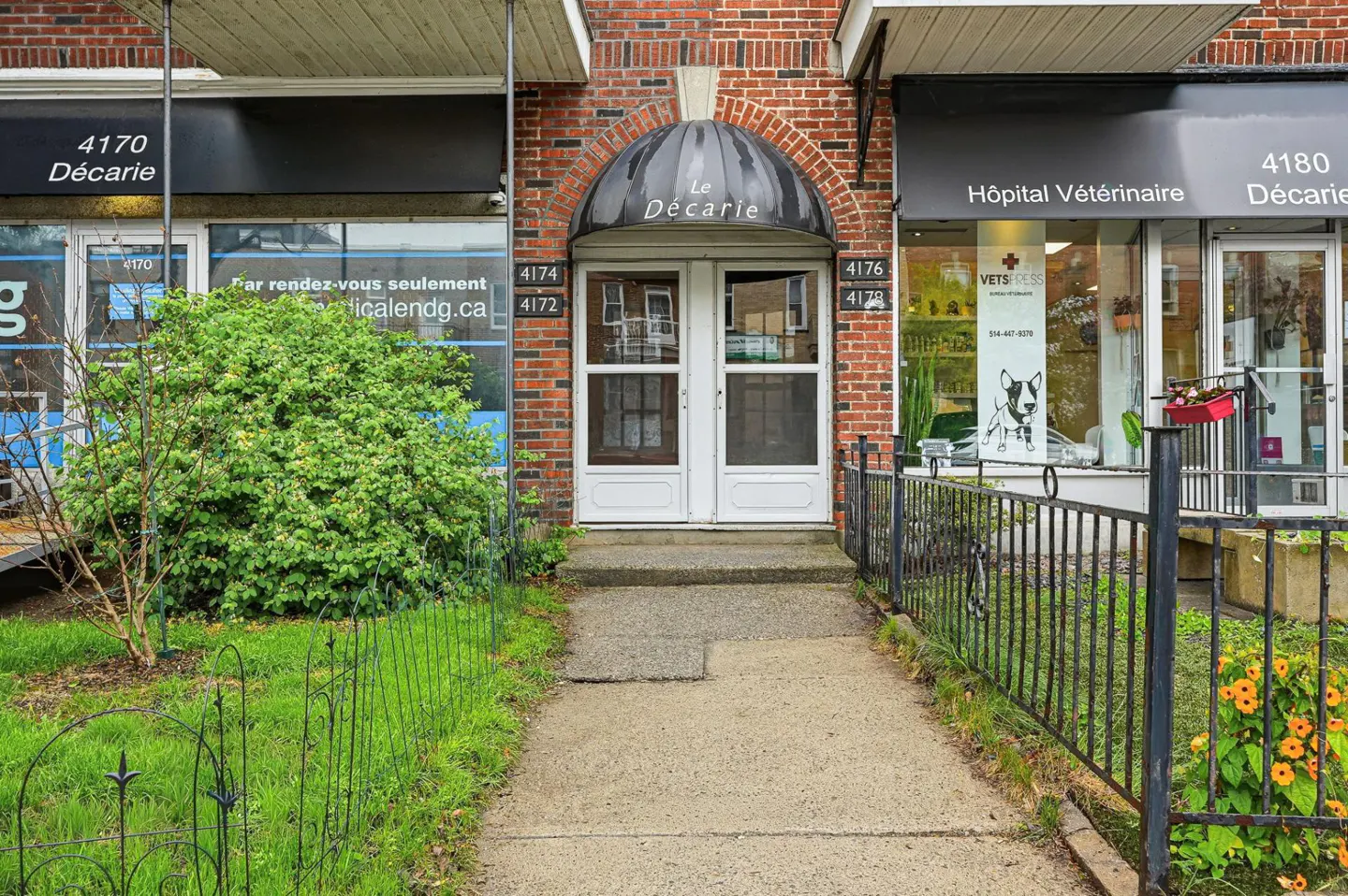 Brick building with "Le Décarie" awning over white double doors. Sidewalk leads to entrance, flanked by green grass and black metal fences.