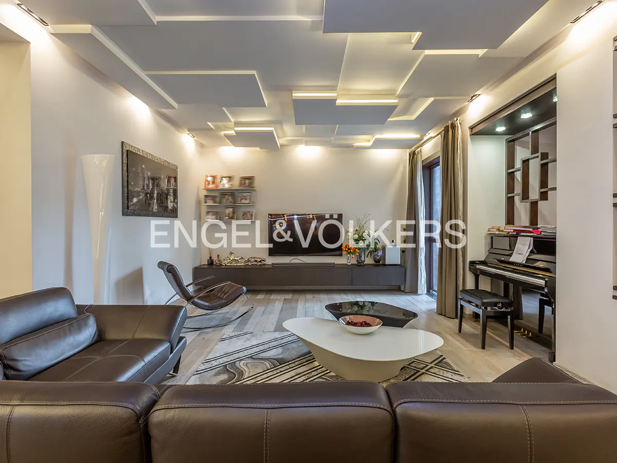 Living room with brown leather sofa, white coffee table, piano, and modern ceiling with recessed lighting. Engel & Volkers logo visible.