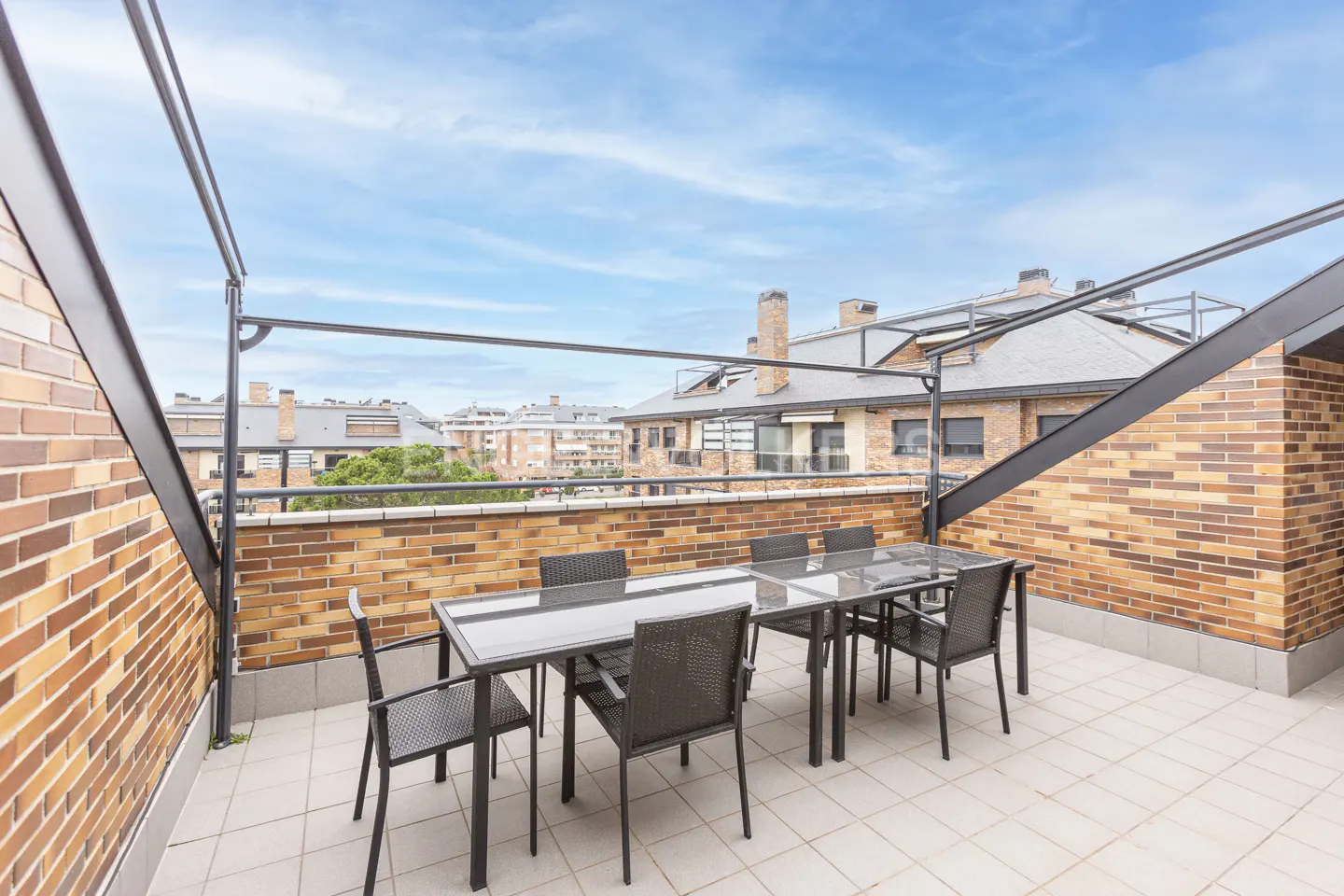 Outdoor patio with a glass table and six chairs, brick walls, and a view of buildings under a blue sky.
