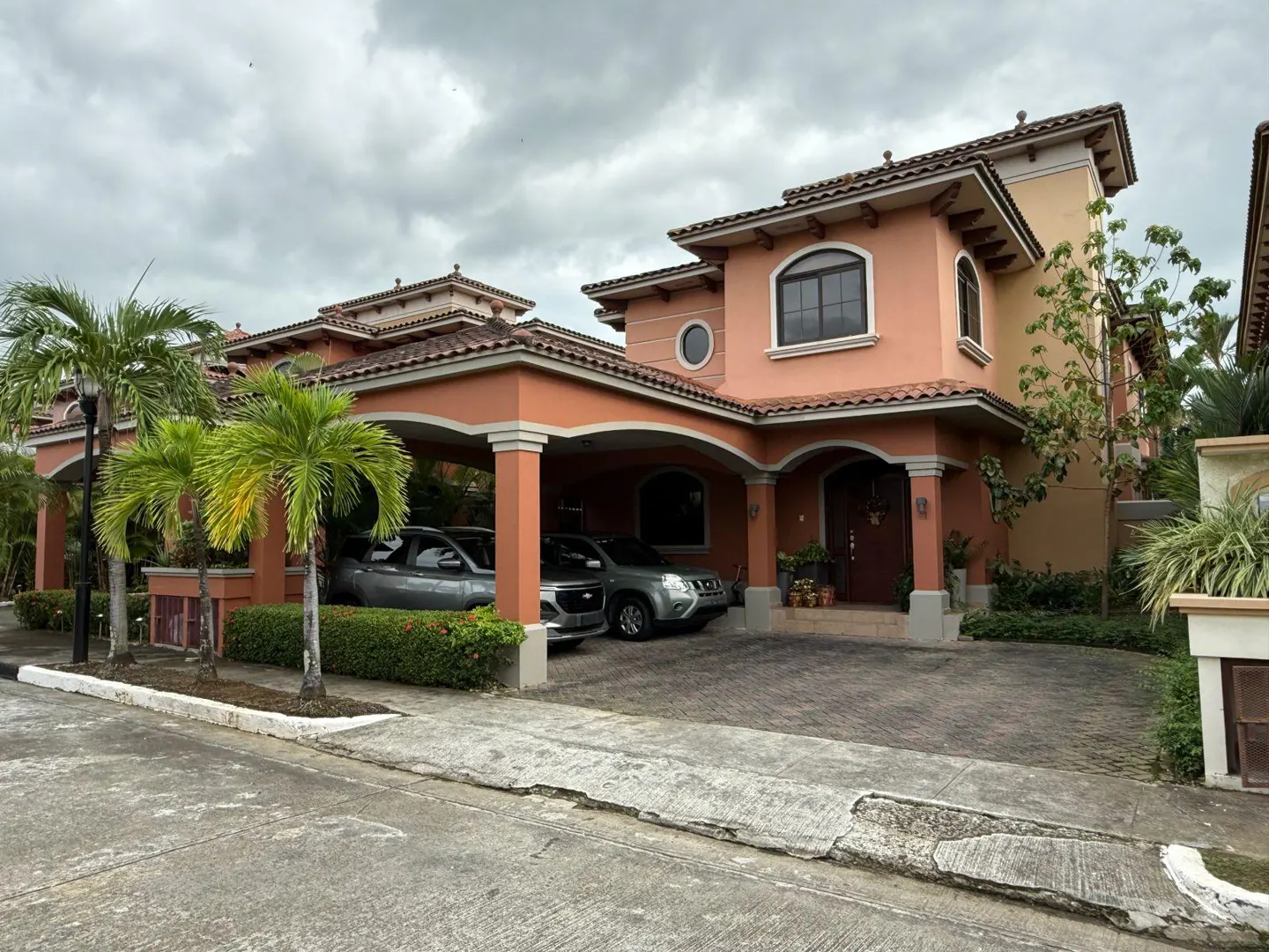 Two-story peach house with a tile roof and a two-car carport. Two cars are parked in the carport. Palm trees in the front yard.