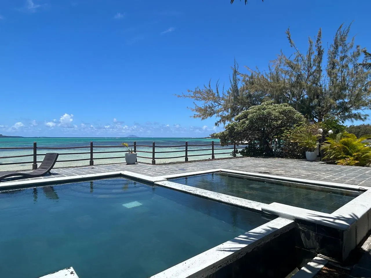 Outdoor pools with ocean view. Blue sky, turquoise water, and a lounge chair by a wooden fence.