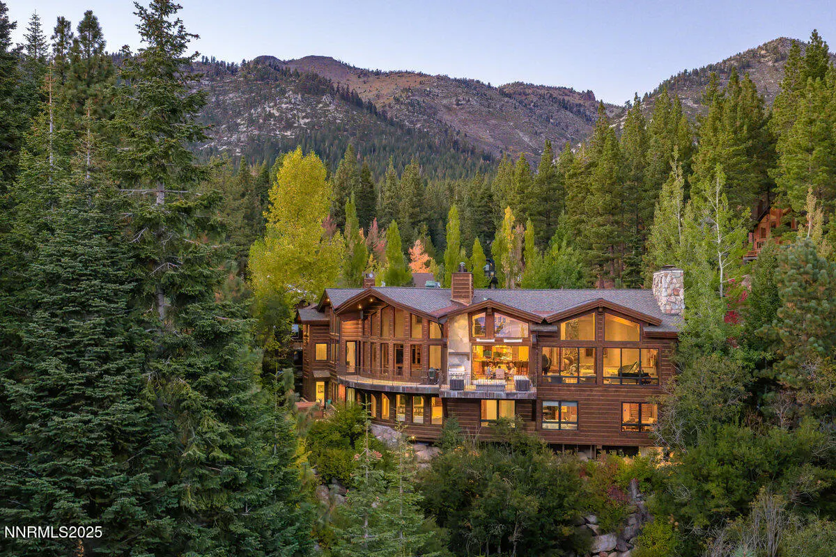 A large, brown, log cabin-style house nestled among green trees and mountains under a blue sky.