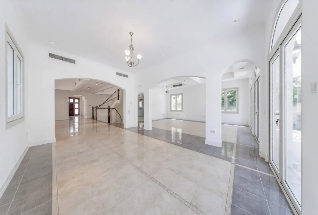 Bright, empty room with white walls, arched doorways, and tile flooring. A staircase is visible in the background. Large windows let in natural light.