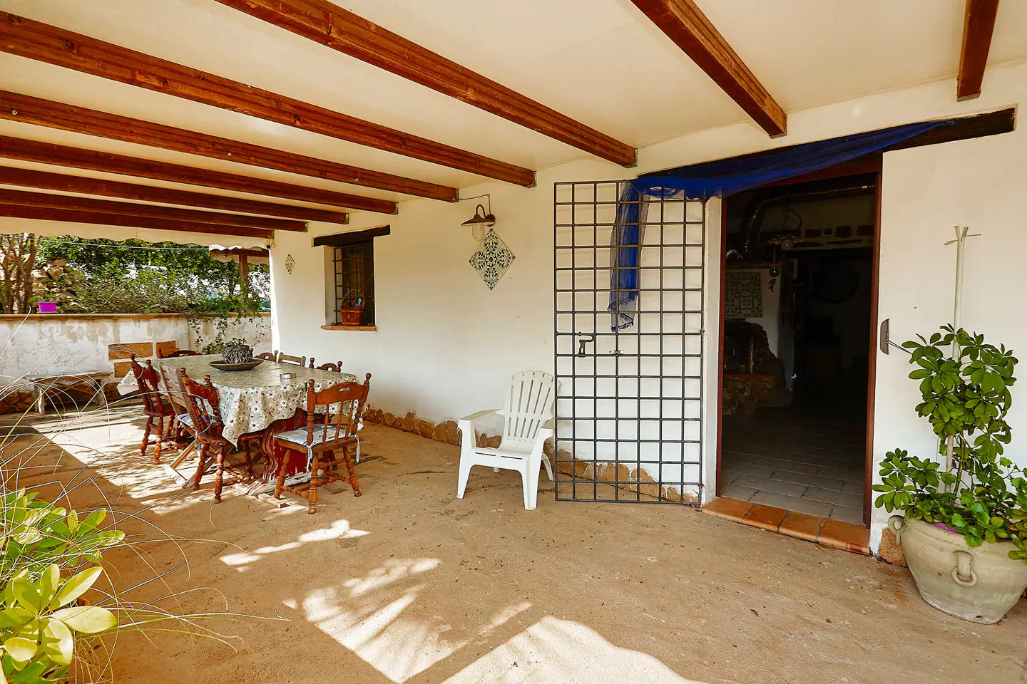 Covered patio with a dining table, chairs, and a white plastic chair. A metal grid stands near an open doorway. A potted plant sits to the right.