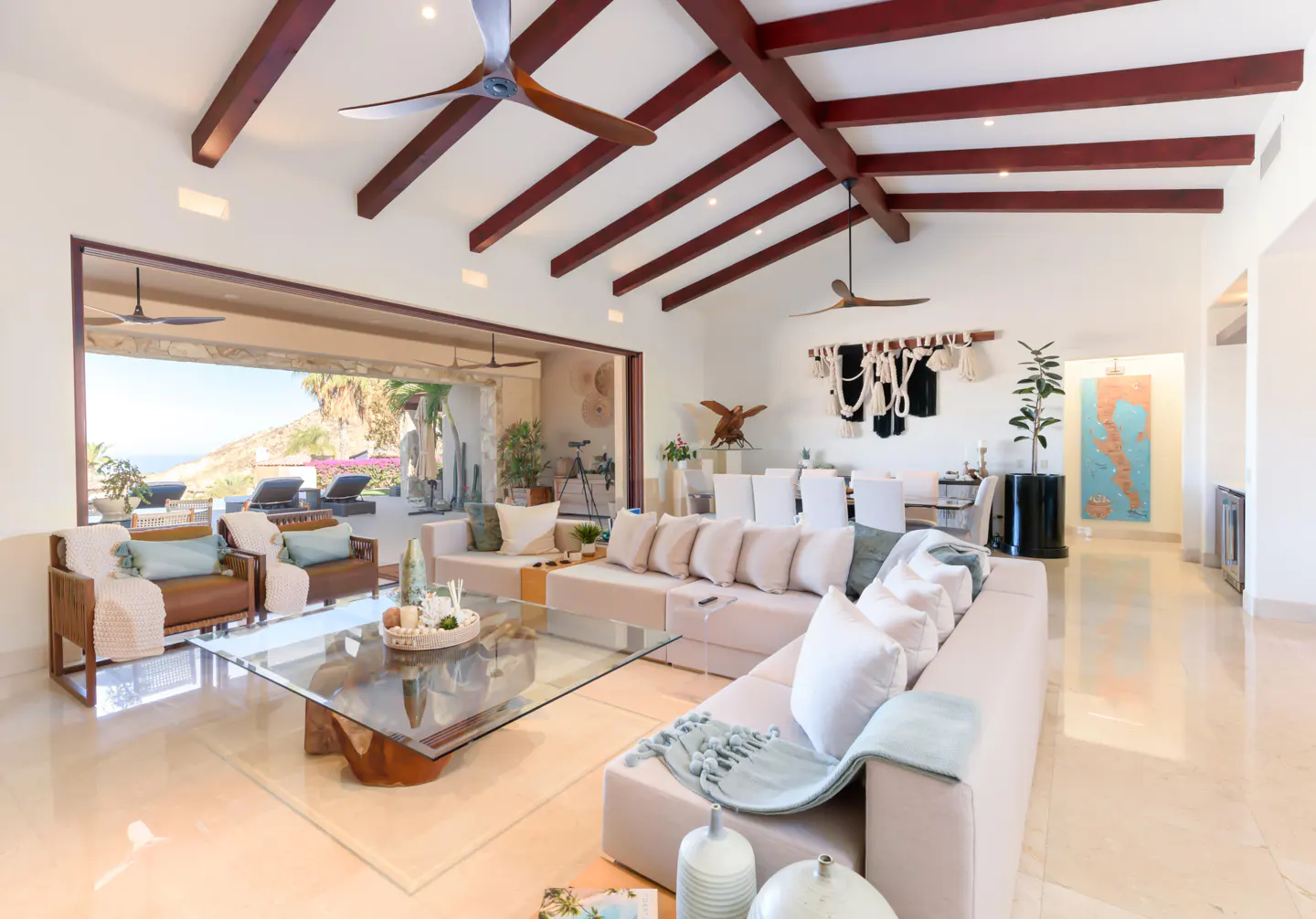 Bright living room with beige sectional, glass table, and ocean view through open doors. Red ceiling beams and ceiling fans.