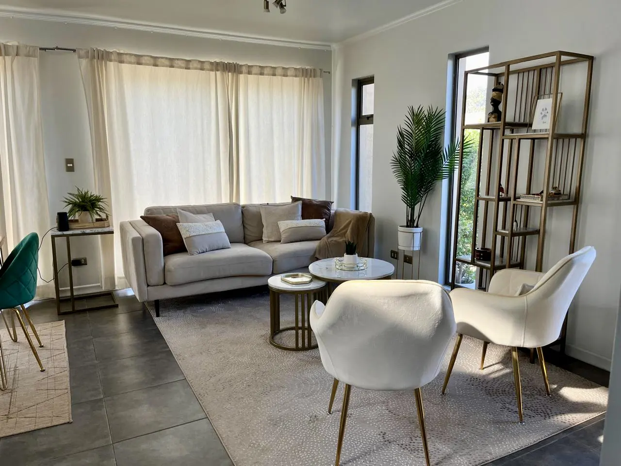 Bright living room with beige sofa, white chairs with gold legs, and a gold bookshelf near a window.