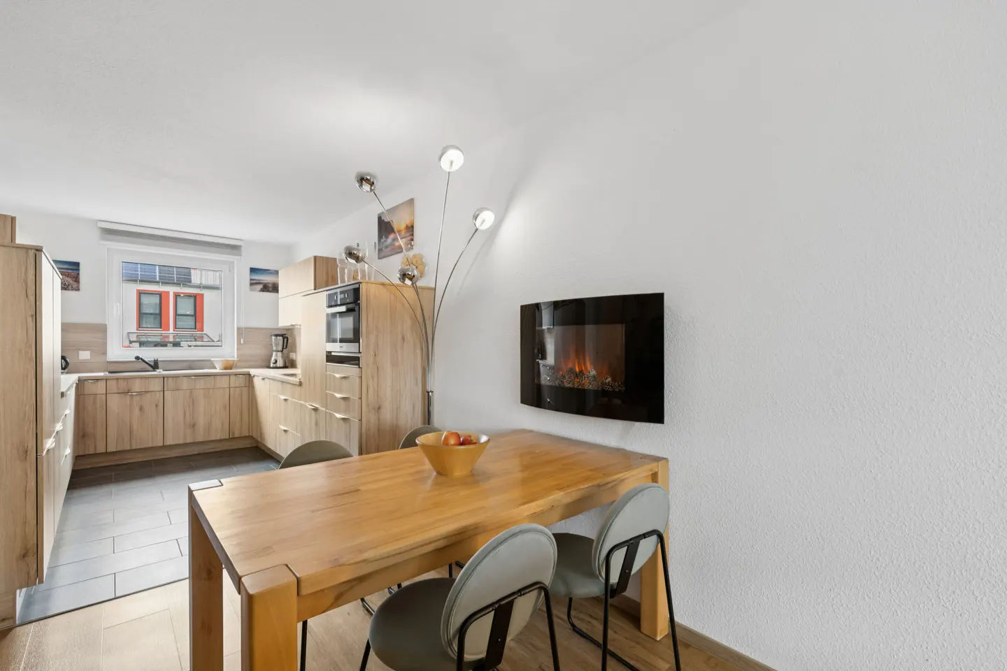 Bright kitchen with light wood cabinets, a dining table with gray chairs, and a black electric fireplace on a white wall.