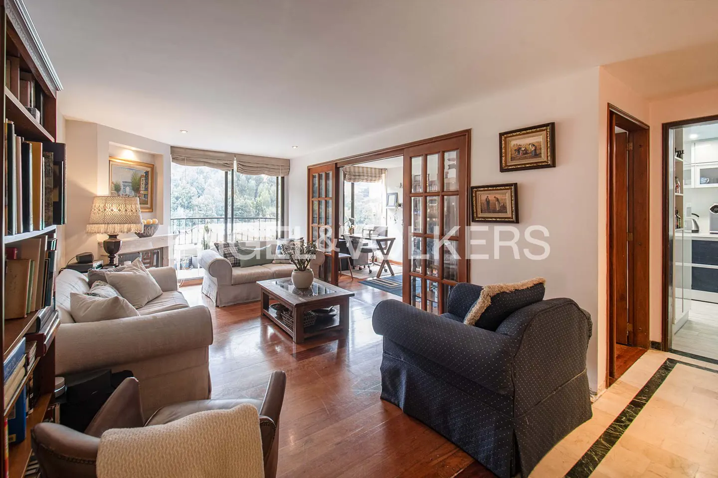 Bright living room with hardwood floors, sofas, a bookshelf, and a view to a balcony through glass doors.