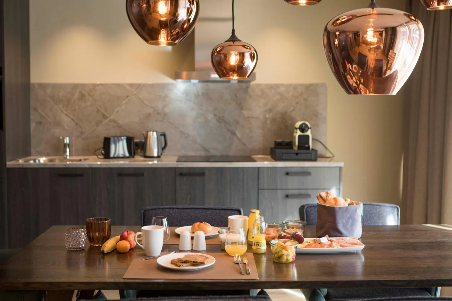 A dining table set for breakfast with toast, fruit, and drinks, under modern copper pendant lights in a modern kitchen.