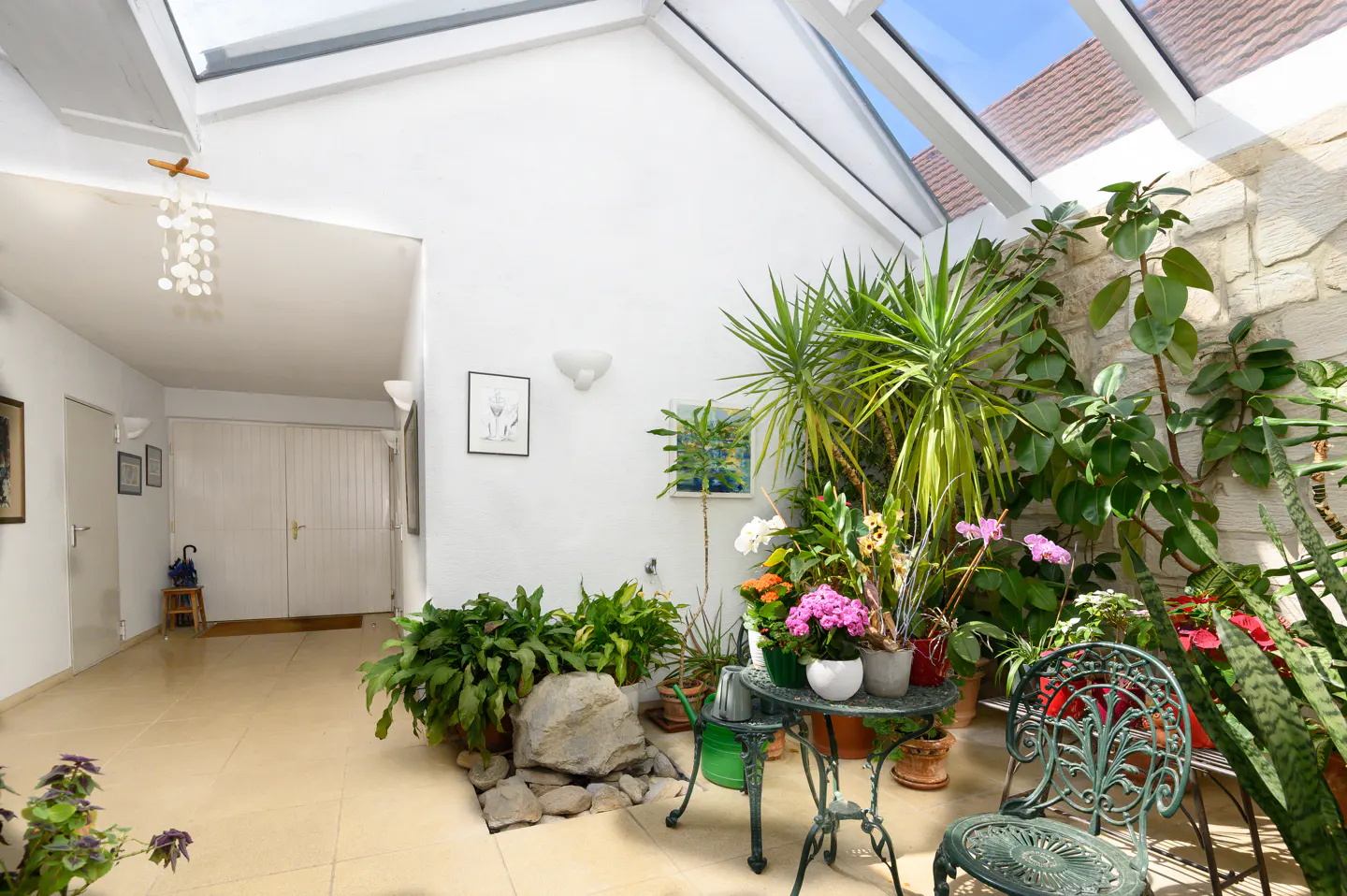Bright sunroom with skylight, white walls, and tile floor. Green plants and flowers surround a wrought iron table and chair set.