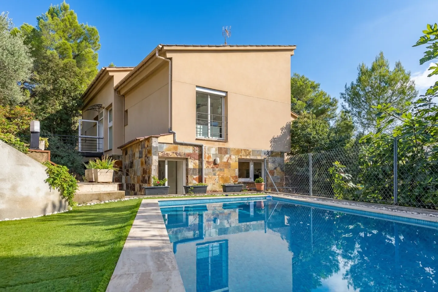 Tan two-story house with a blue tiled pool in the foreground and green trees in the background.