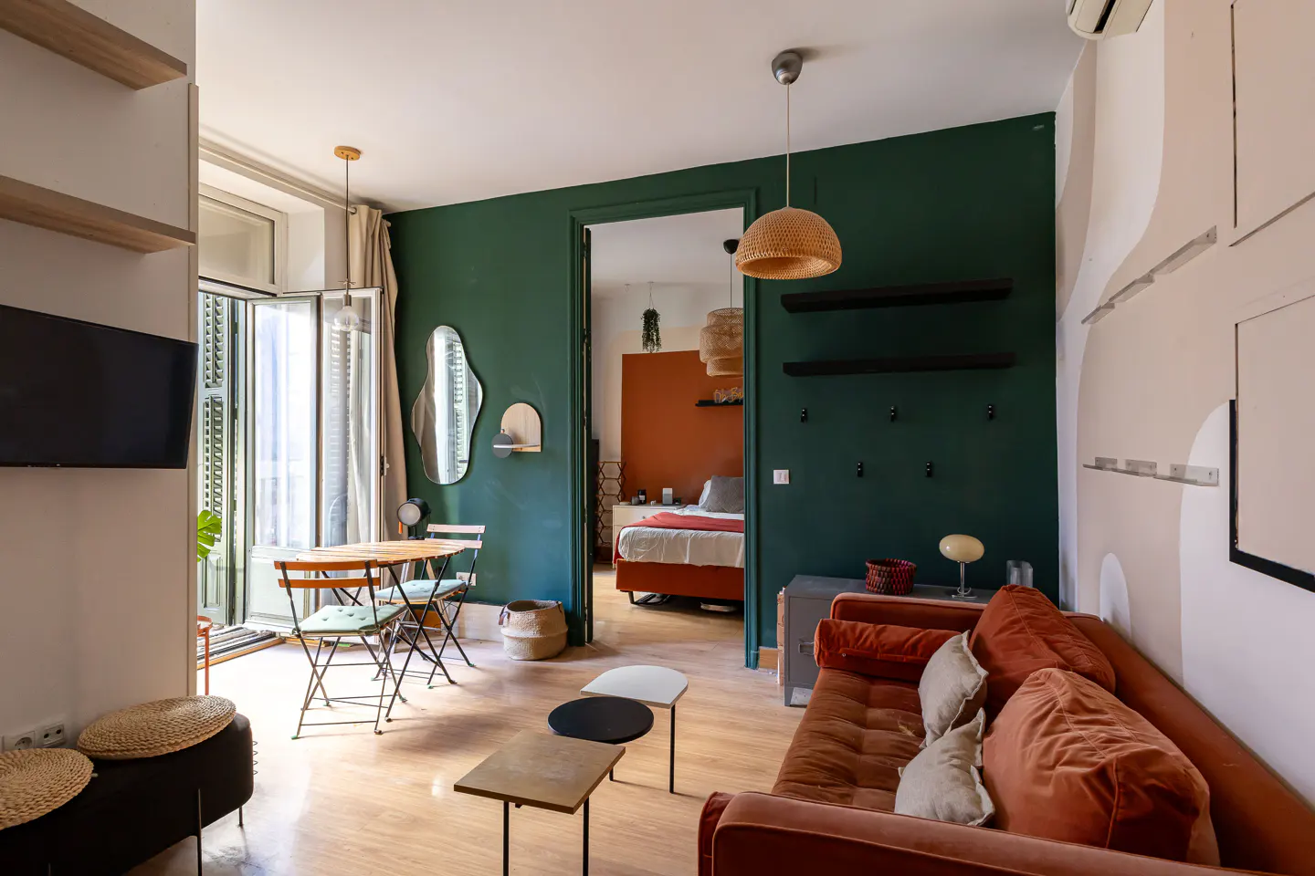 A cozy apartment interior with green walls, a rust-colored sofa, and a view into the bedroom. A small table and chairs are near the balcony.