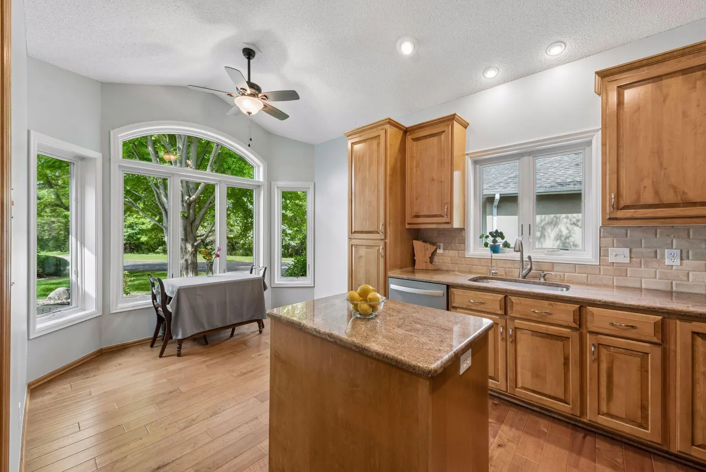 Kitchen with wood cabinets, granite island, hardwood floors, and a dining nook with large windows overlooking greenery.