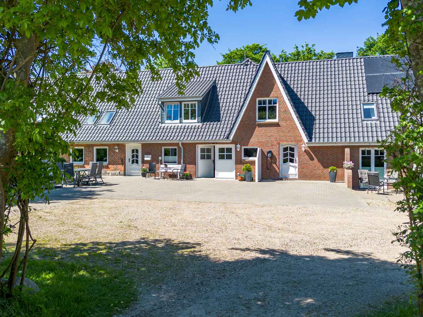 A red brick house with a dark tiled roof, white doors, and a paved patio area framed by green trees on a sunny day.