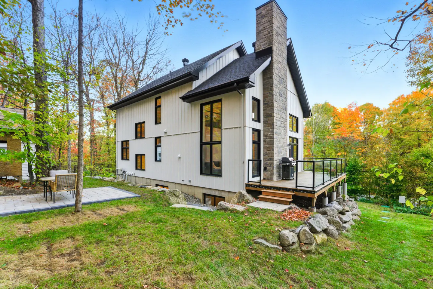 Modern white house with black trim, stone chimney, and deck overlooking a green lawn and colorful autumn trees.