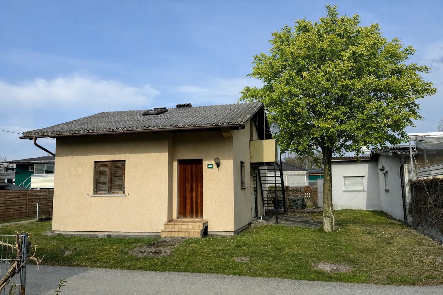 A one-story beige house with a brown door and a gray roof, next to a green tree.