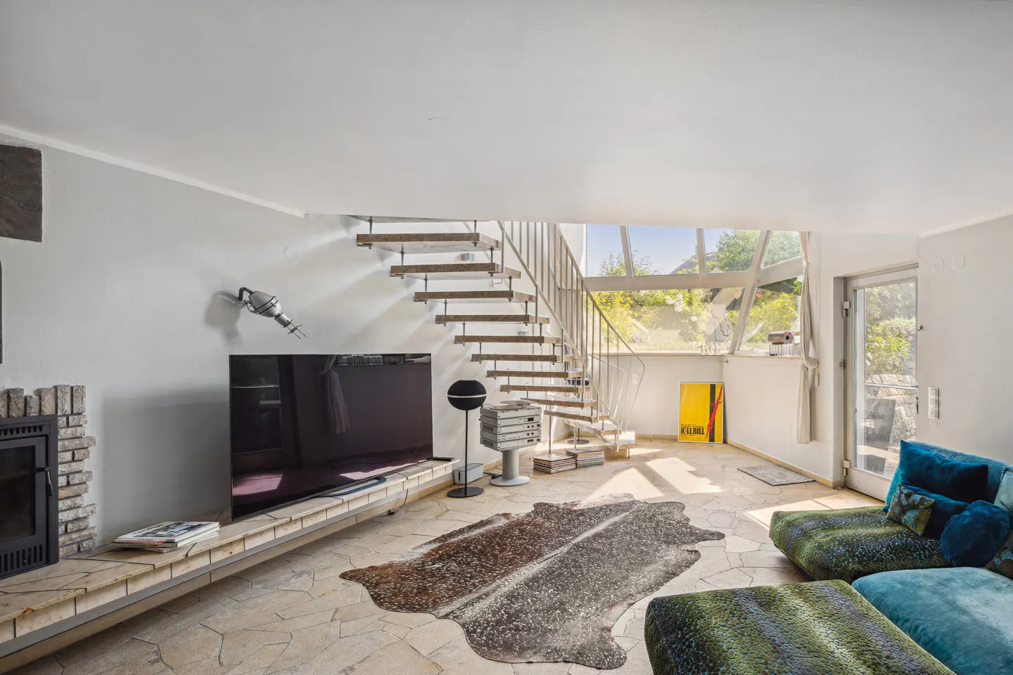 Living room with stone floors, a cowhide rug, and a blue and green patterned sofa. A modern staircase leads to a window with a view of greenery.