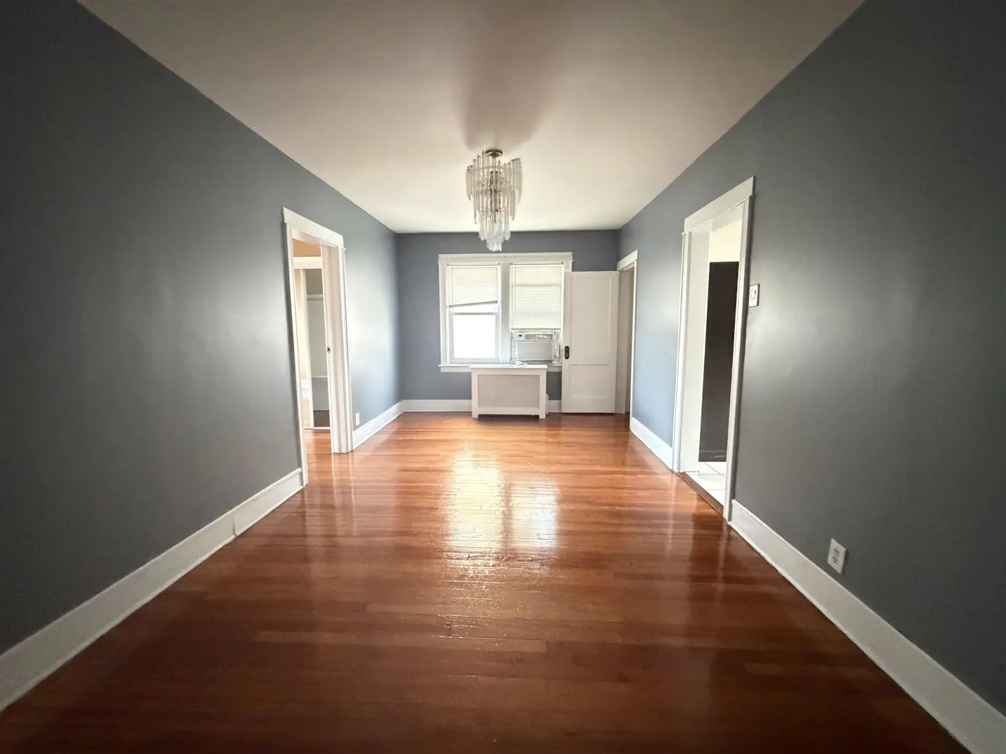 Empty room with blue-grey walls, polished hardwood floors, a crystal chandelier, and a window with a white radiator below.