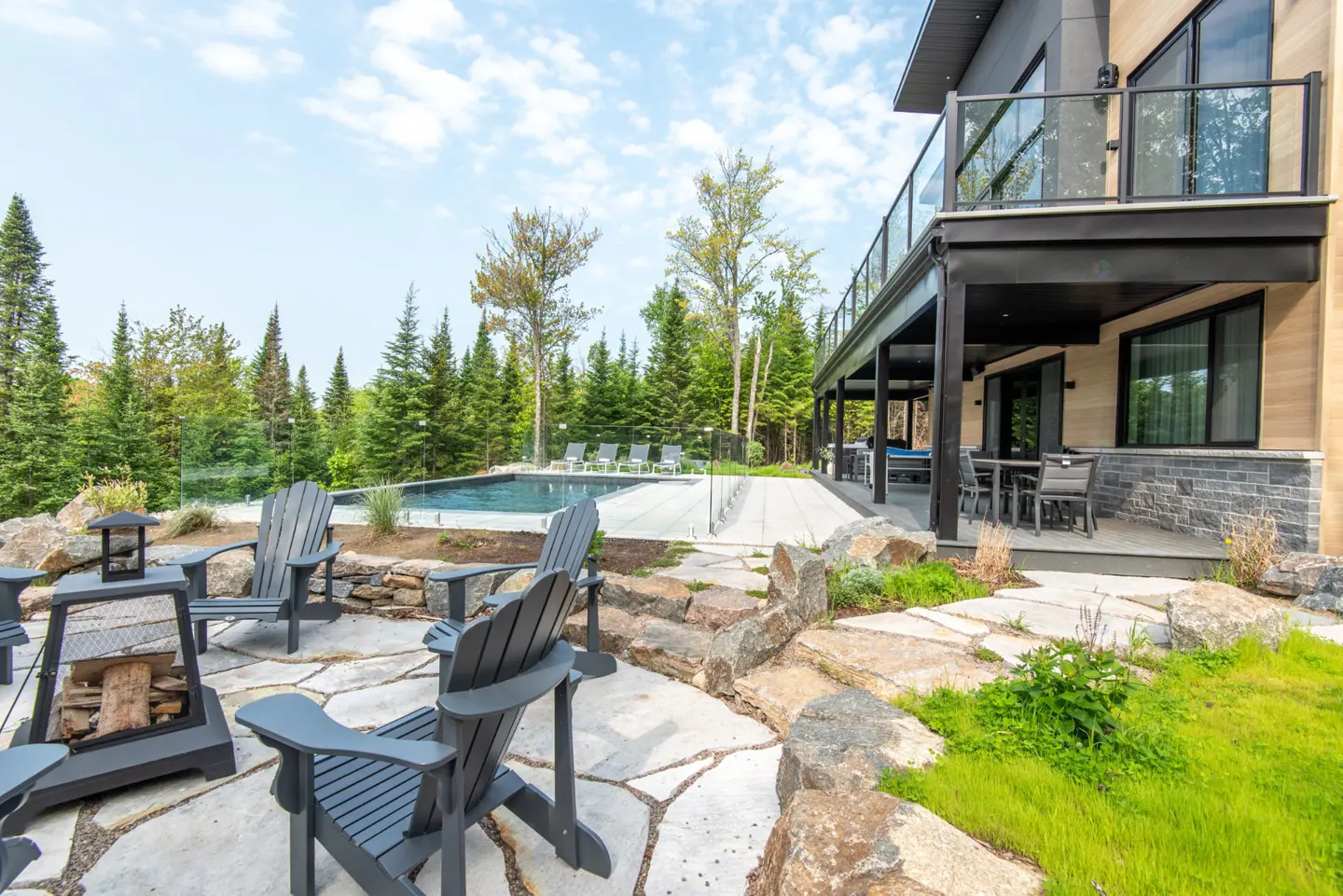 Outdoor patio with gray Adirondack chairs around a fire pit, pool, and modern house with a balcony in a wooded area.