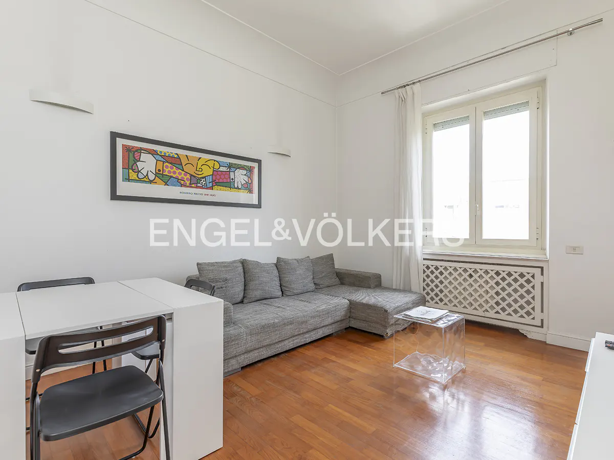 Bright living room with hardwood floors, a gray sectional sofa, a white table with black chairs, and a colorful art print on the wall.