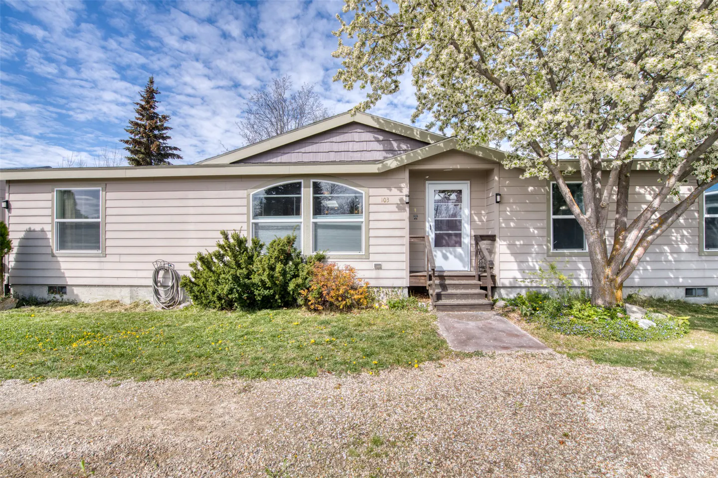 Tan single-story home with a white door, small porch, and a flowering tree in the front yard. Blue sky with clouds above.