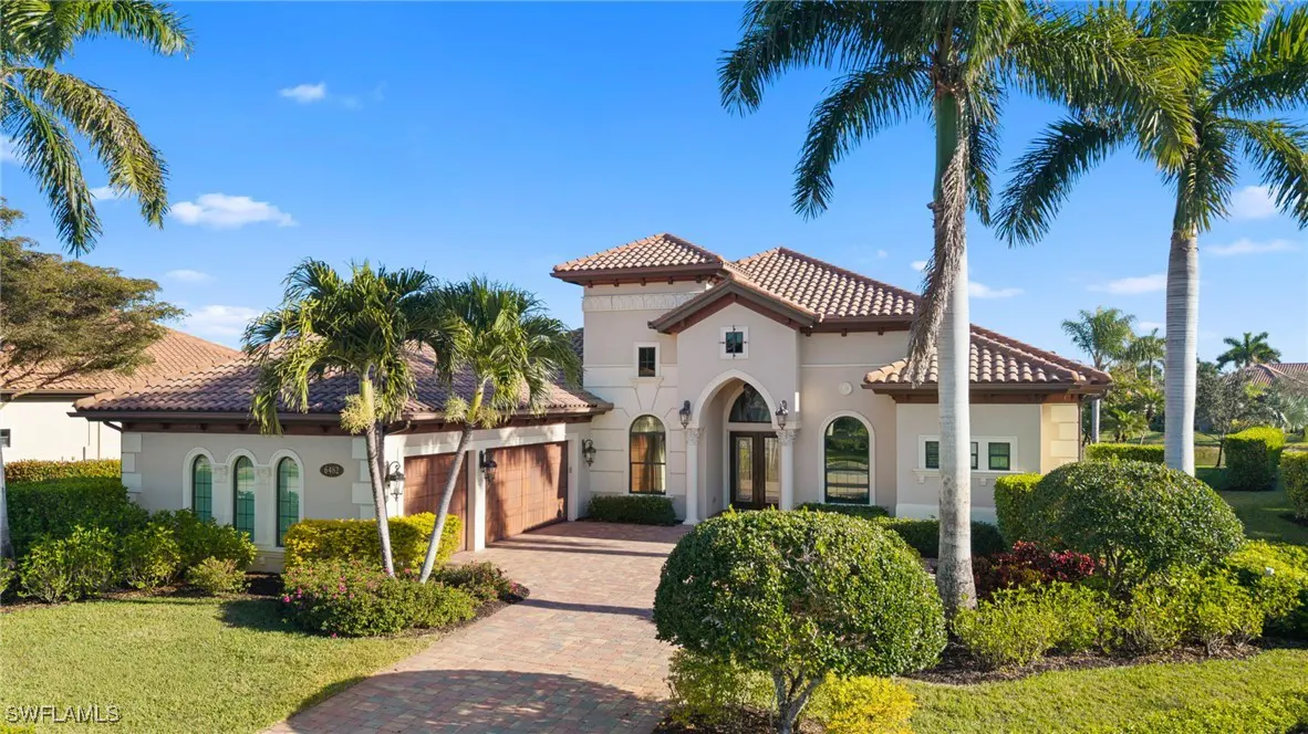 Beige two-story house with a red tile roof, palm trees, and a brick driveway on a sunny day.