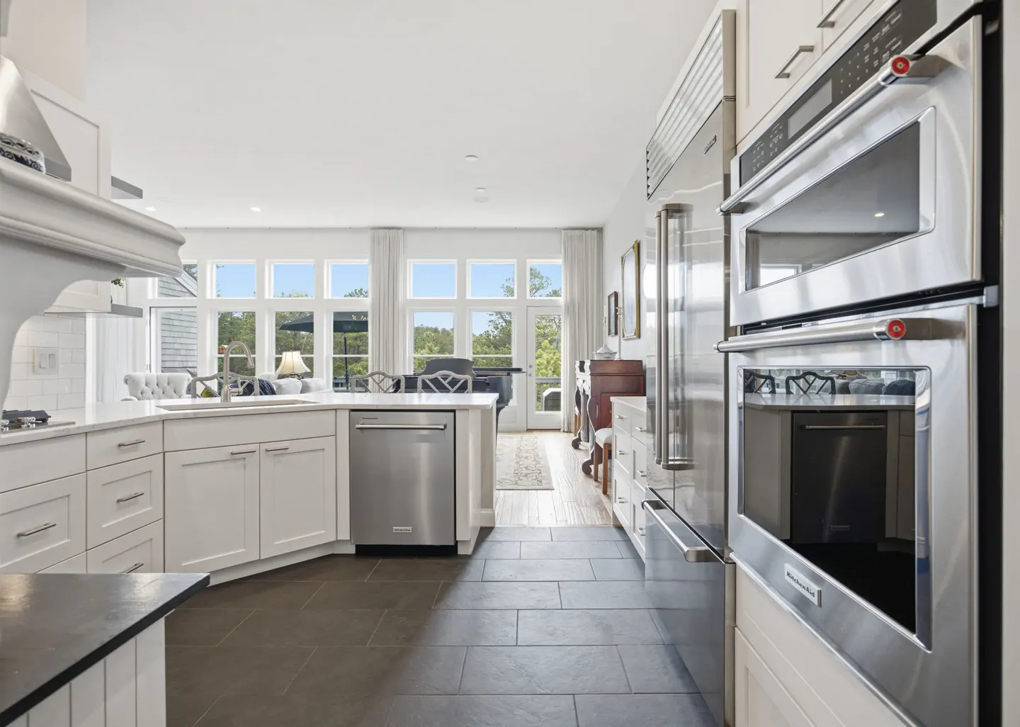 Bright, modern kitchen with white cabinets, stainless steel appliances, and dark tile floors. Large windows offer a view of trees and outdoor seating.