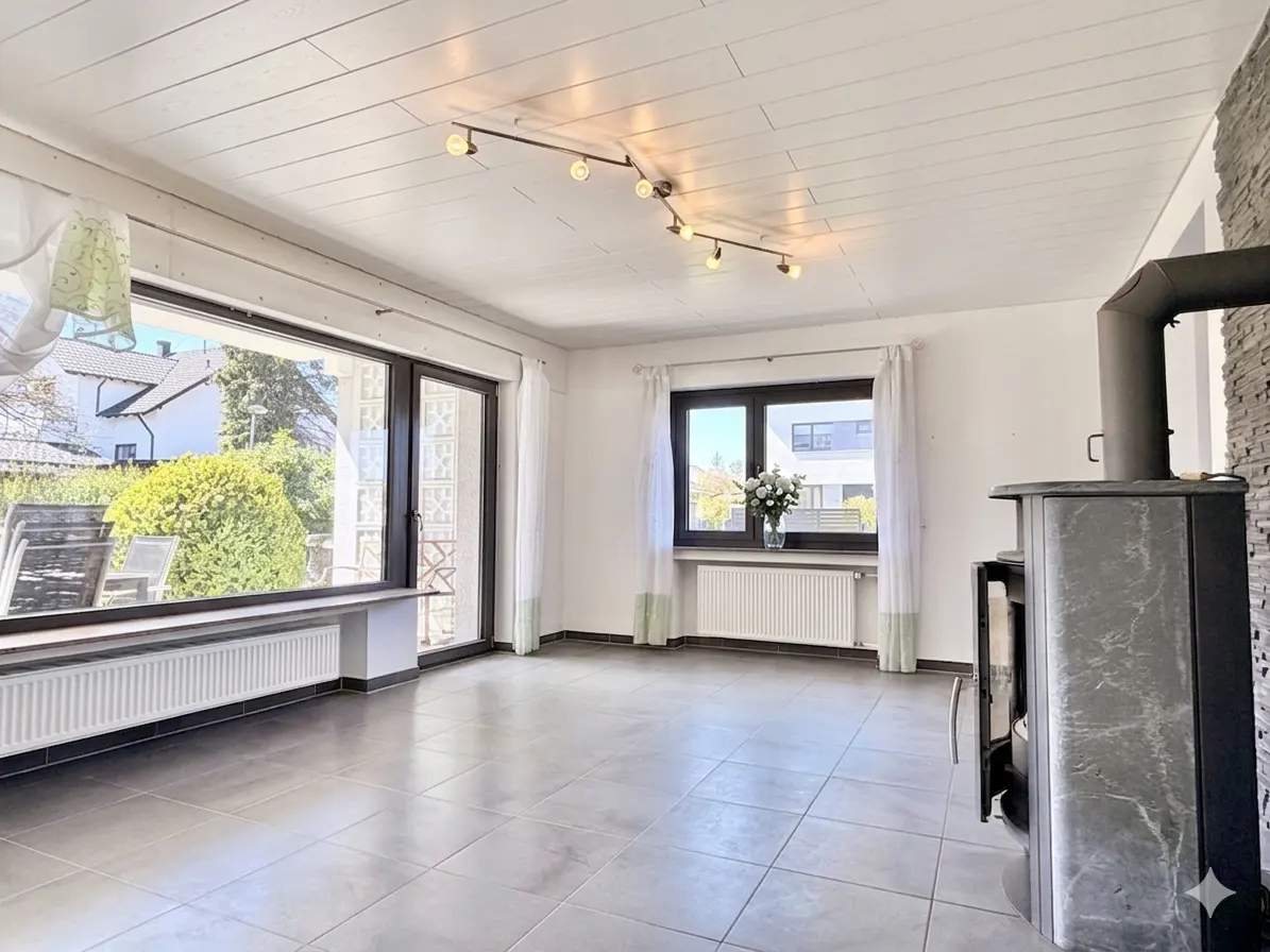 Bright living room with gray tile floor, white walls, and a wood-burning stove. Large windows offer views of the outside.