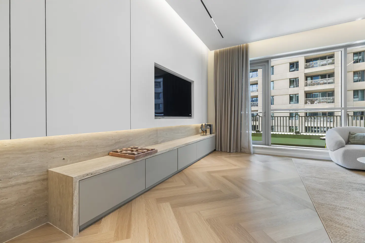 Bright living room with herringbone wood floors, a gray cabinet, and a wall-mounted TV. Large windows overlook a building and balcony.