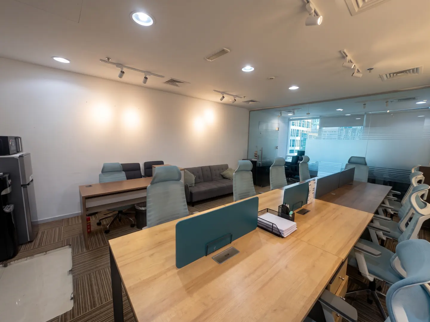 Bright office space with light wood desks, blue chairs, and a gray sofa. Glass-walled conference room in the background.