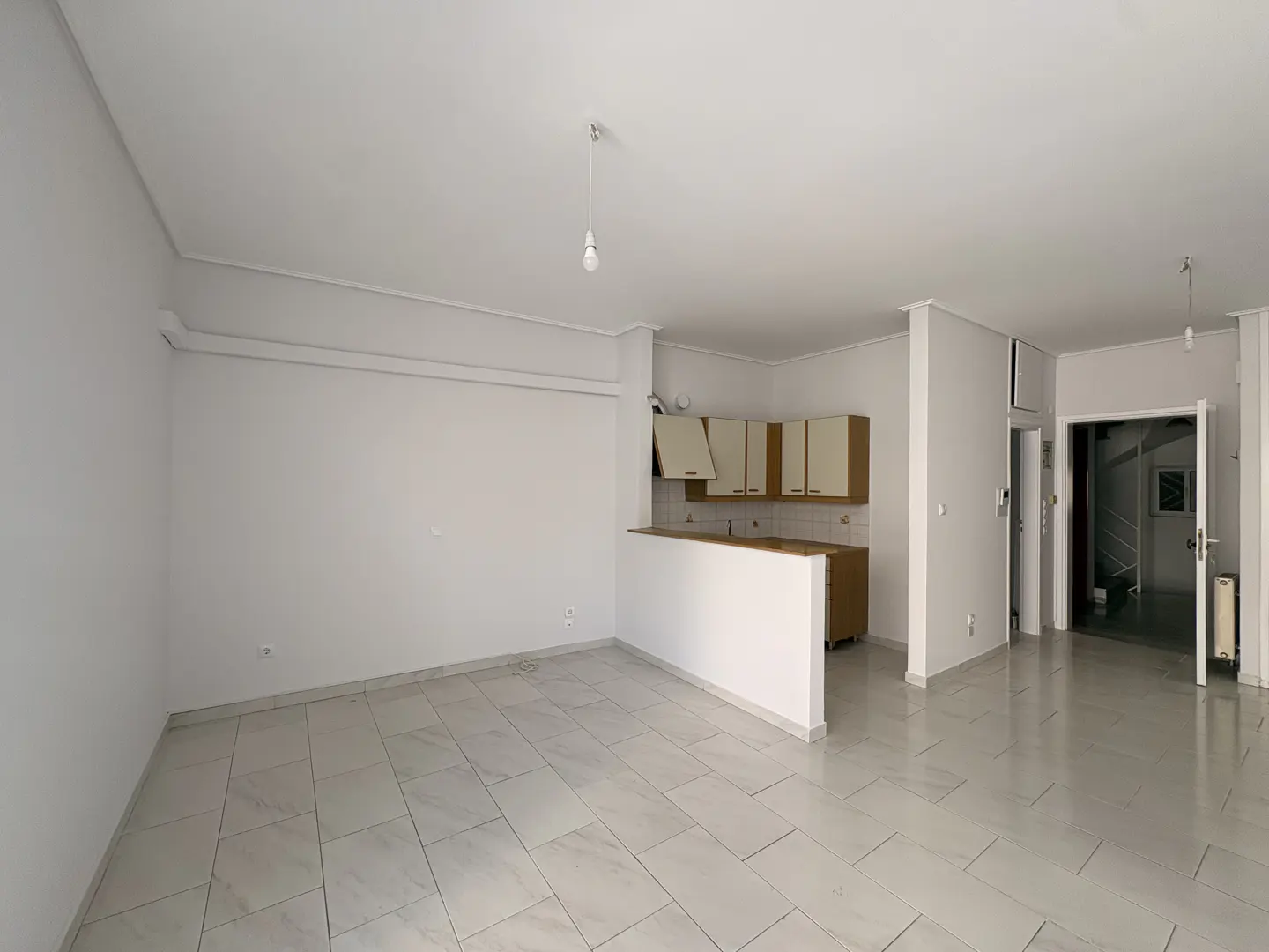 Empty living room with white walls, light tile floors, and an open kitchen area with wood-toned cabinets.