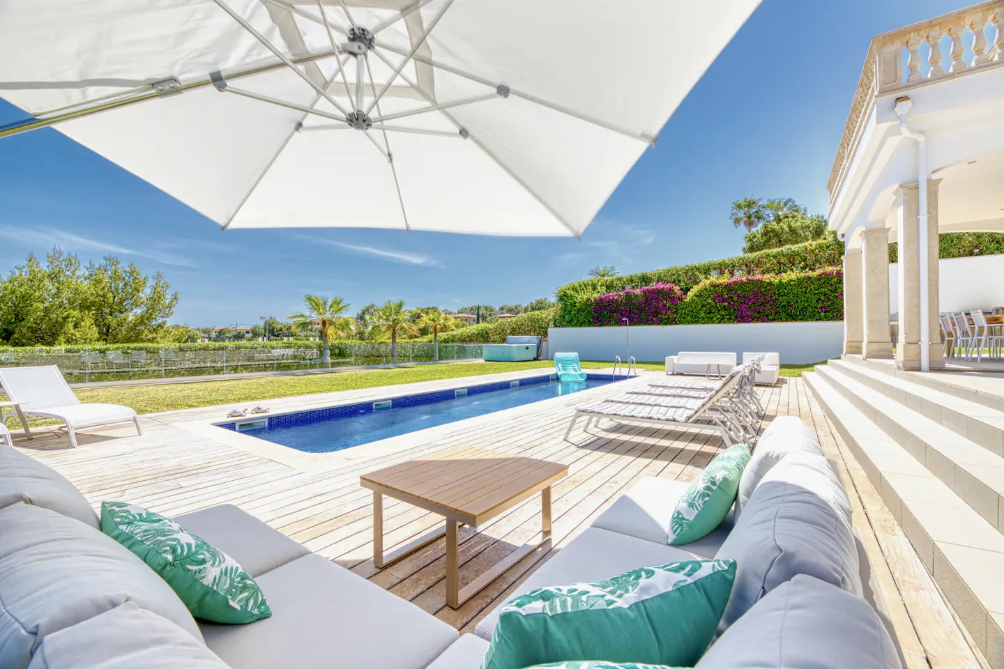 Luxury patio with a swimming pool, white lounge furniture, green palm trees, and a large white umbrella under a blue sky.