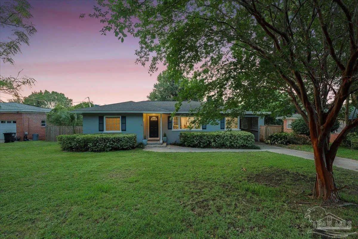 A single-story, light blue house with dark shutters and a black door sits on a green lawn at dusk. A large tree is on the right.