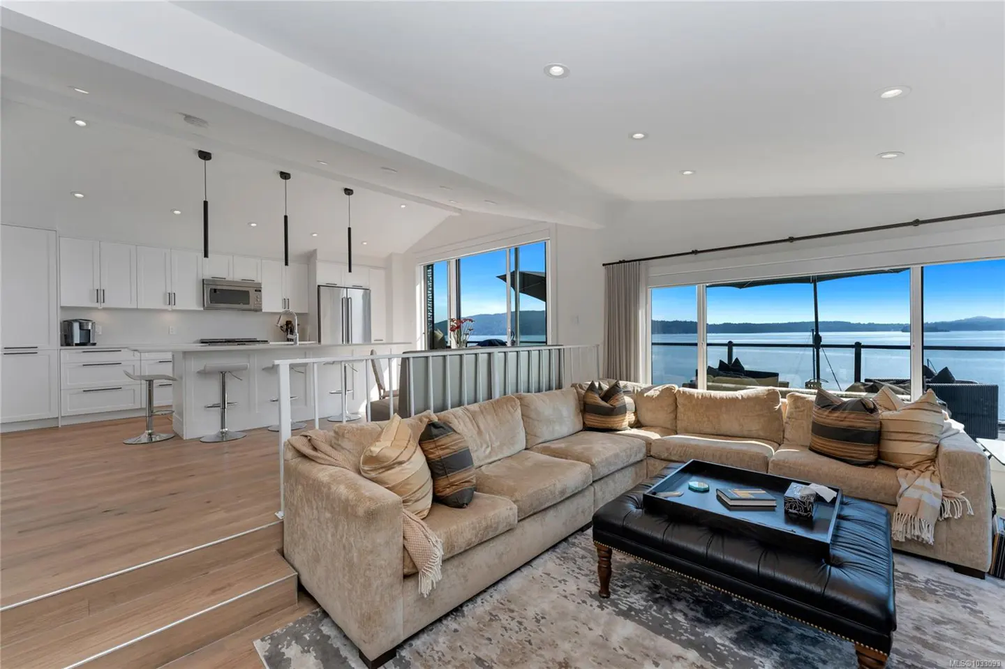 Bright, open-concept living space with a beige sectional sofa, black ottoman, and a kitchen with white cabinets overlooking a lake.