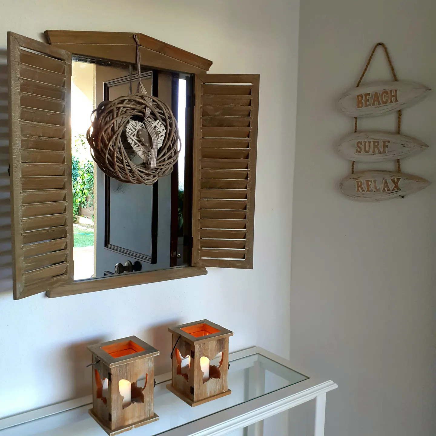 Interior shot of a white wall with a mirror framed by wooden shutters, two candle lanterns, and a beach-themed sign.