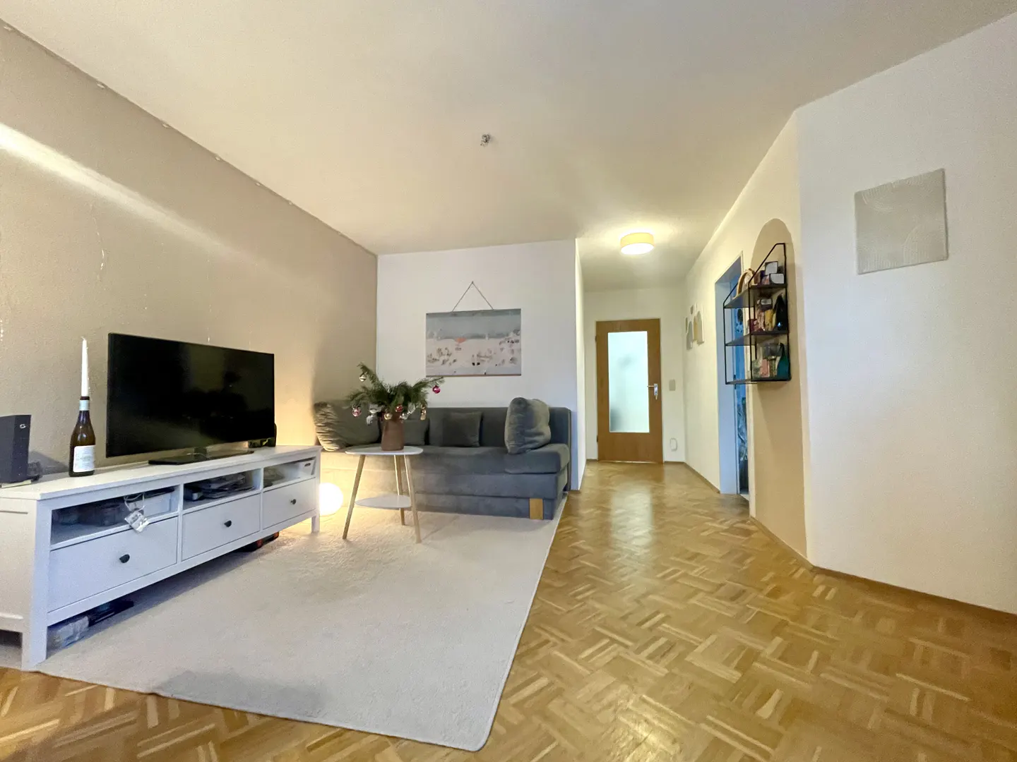 Living room with a gray sofa, white TV stand, and herringbone wood floors. A white rug covers the floor in front of the sofa.