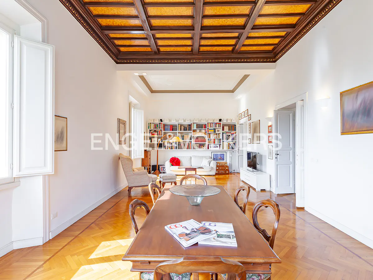 A bright, spacious living room with a wooden dining table, chairs, and a bookshelf in the background.