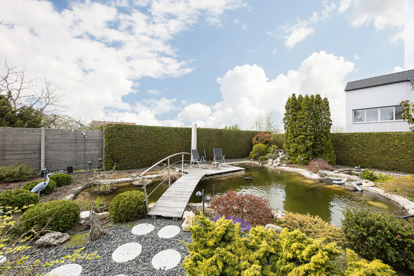 Landscaped backyard with a pond, wooden bridge, and two chairs on a small deck. Green hedges and a blue sky with clouds in the background.