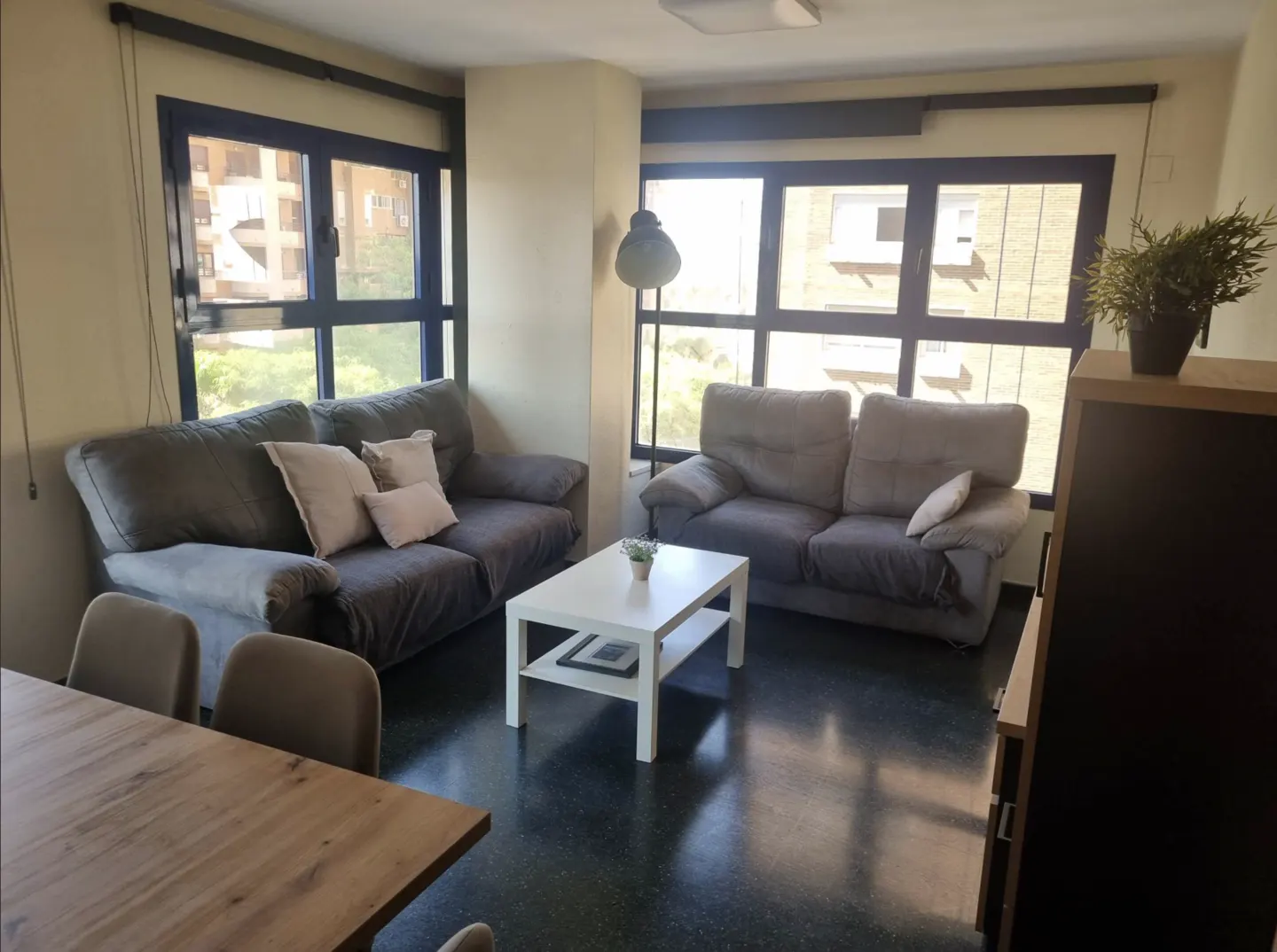 Living room with two grey sofas, a white coffee table, and a wooden dining table, set against large windows.