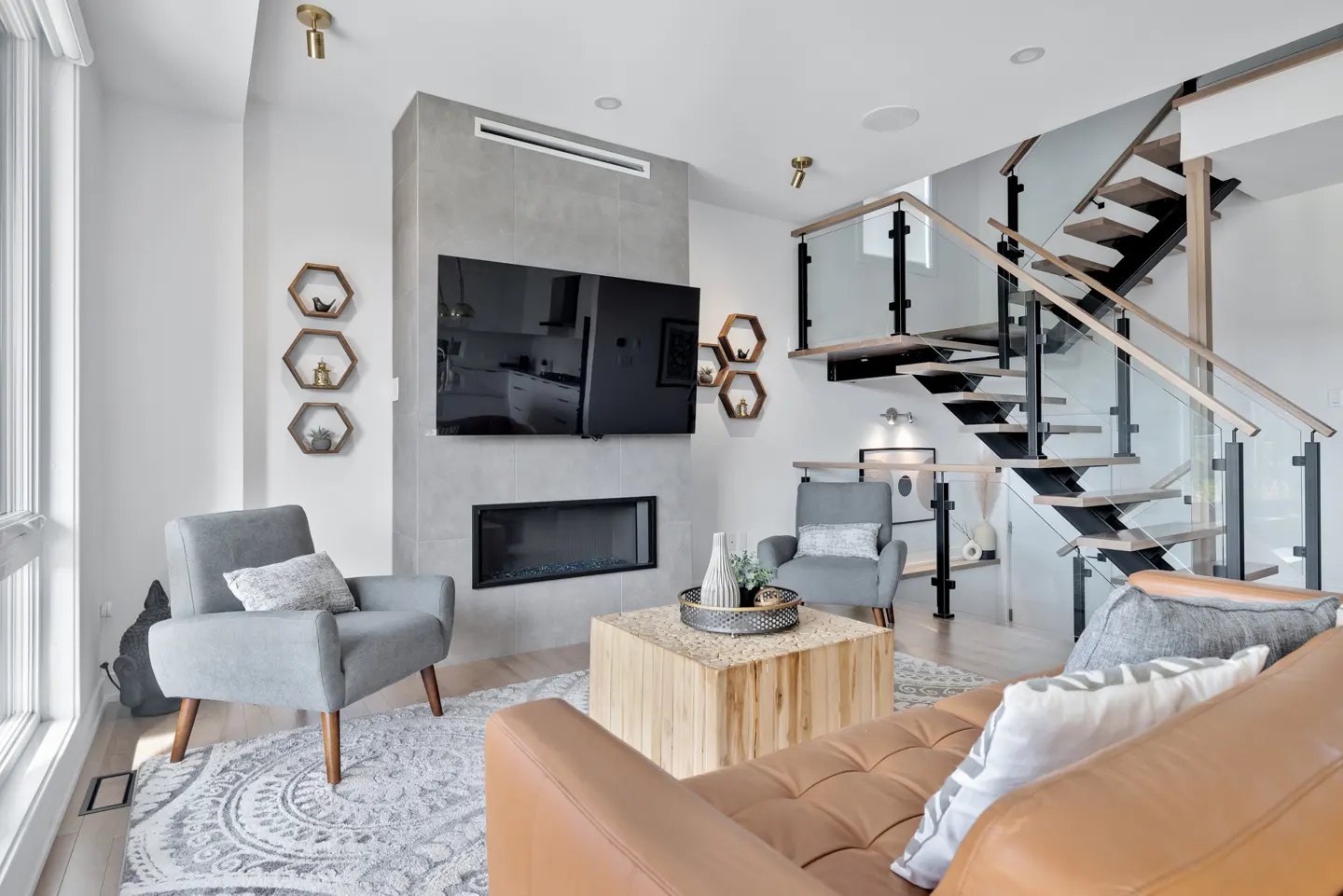 Bright living room with a brown leather sofa, gray chairs, a fireplace, and a staircase with glass railings.