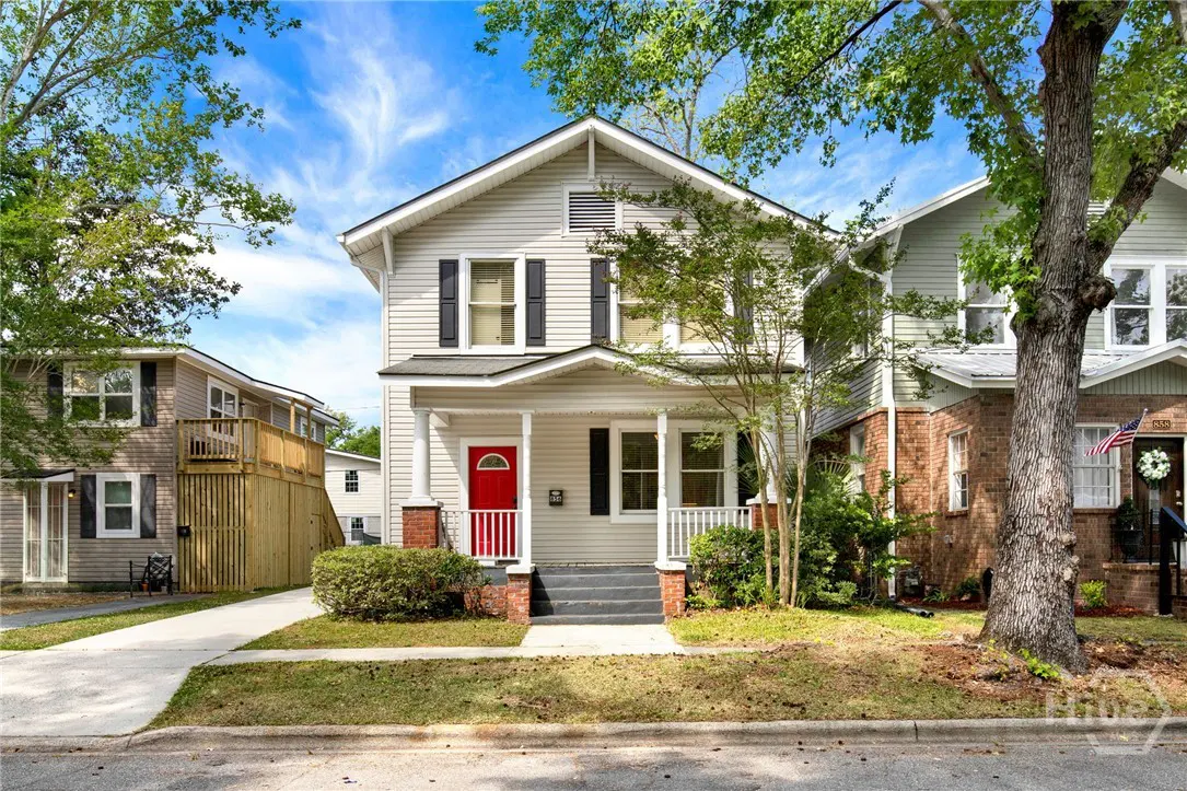 Two-story house with gray siding, black shutters, and a bright red front door. A small porch with white railings and brick columns. Green trees and lawn.