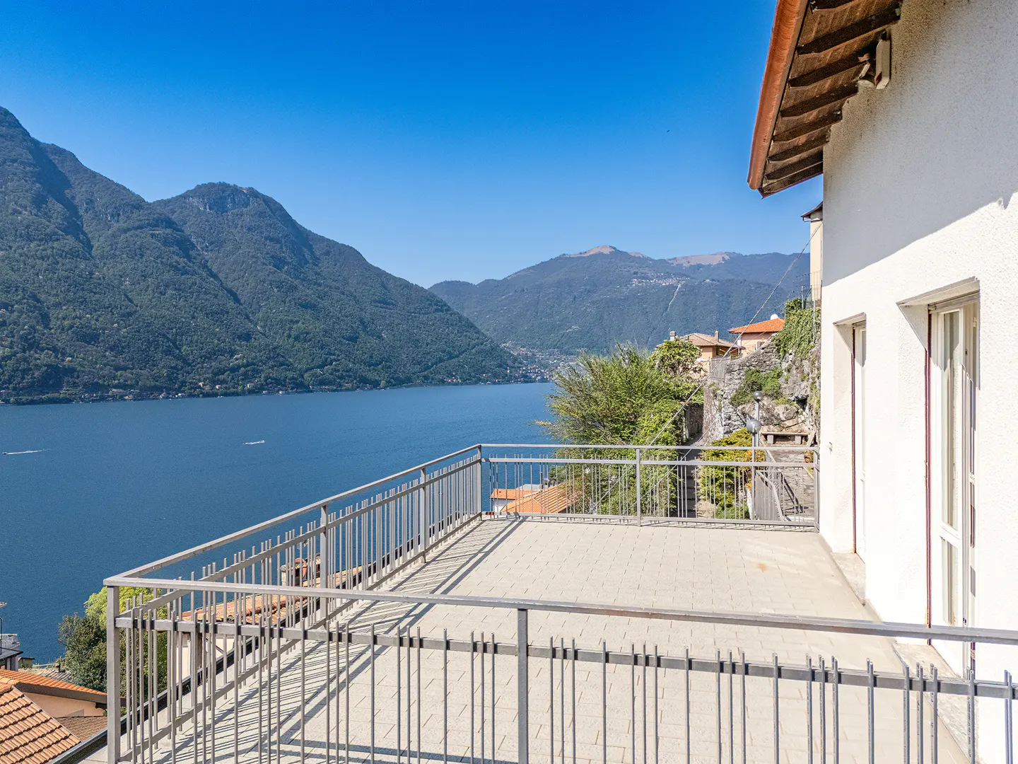 Balcony view of Lake Como, Italy. Metal railing overlooks blue water and green mountains under a clear blue sky. White building on the right.
