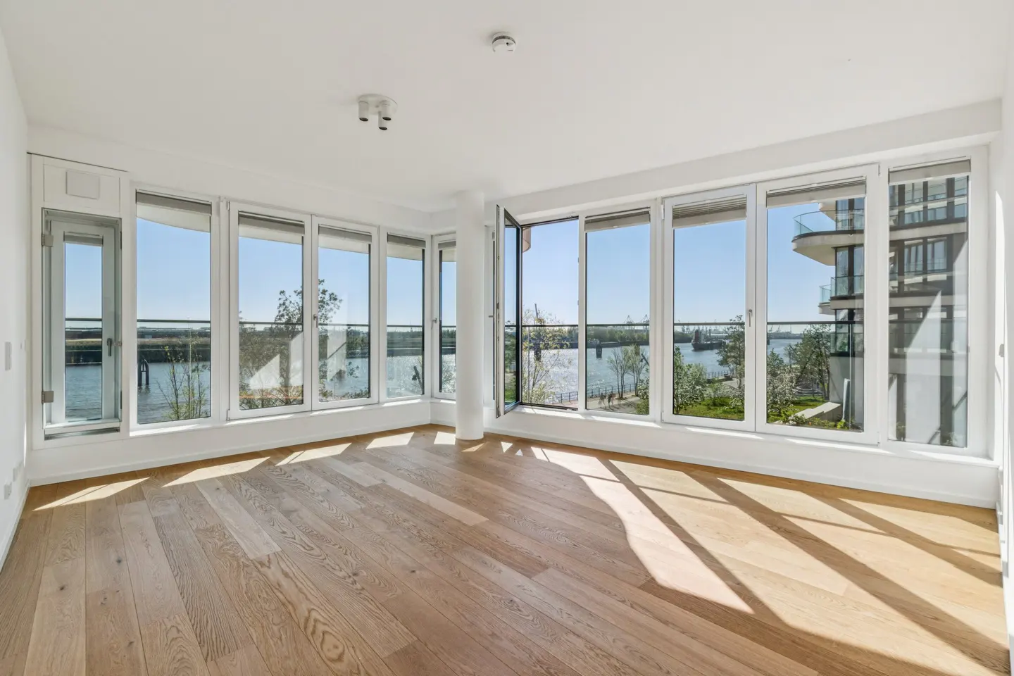 Bright, empty room with light wood floors and white walls. Large windows offer a view of a river and city buildings.