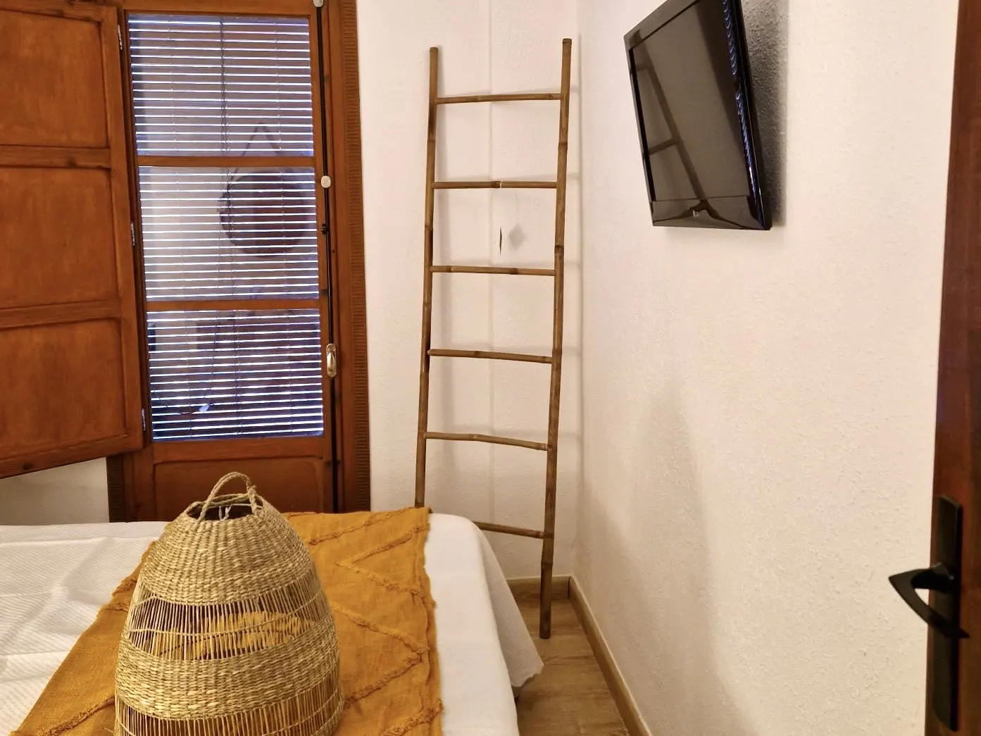 Bedroom interior with a bed, woven basket, yellow throw, wooden ladder, TV, and a window with blinds.